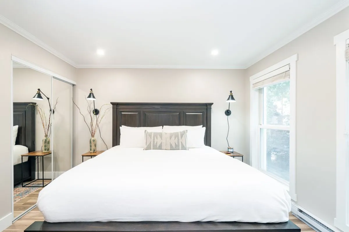 A sunlit bedroom featuring a large bed with decorative pillows, crisp linens, and nightstands on either side. The mirrored closet and soothing color palette create a sense of space and comfort, ideal for unwinding after a busy day. The natural light streaming in highlights the room's serenity.