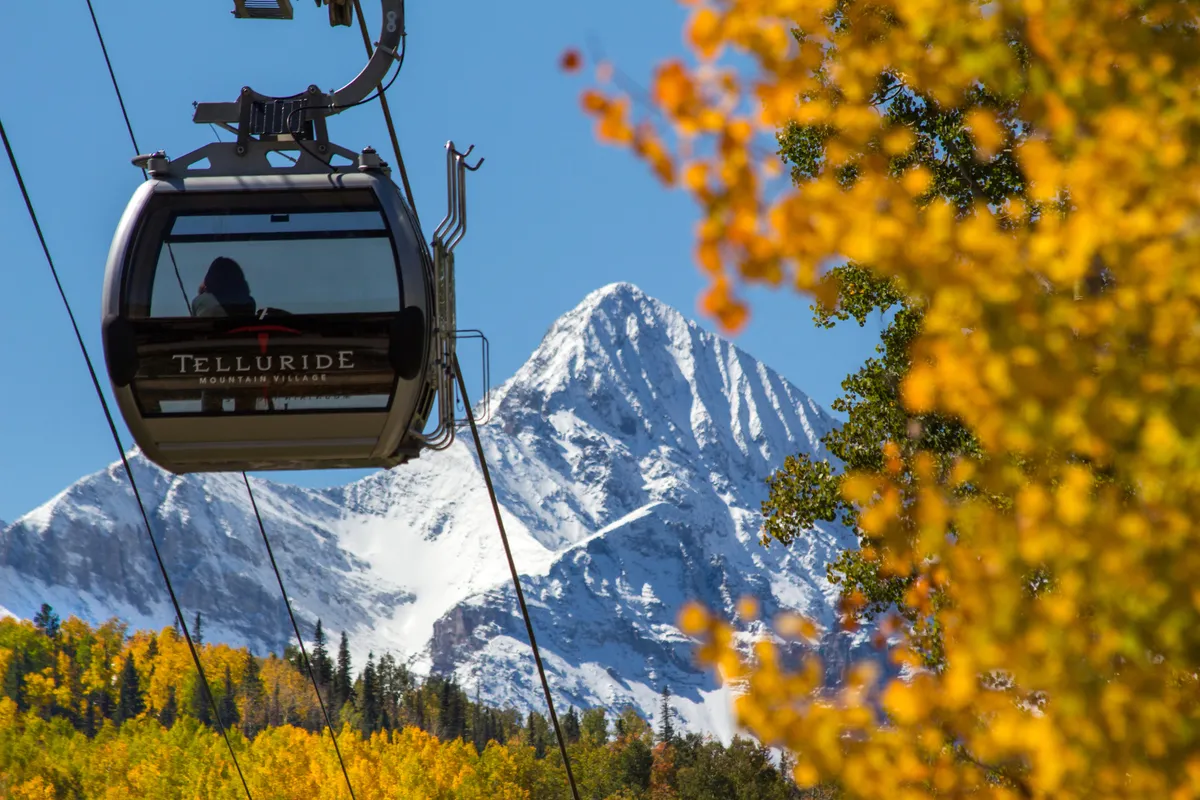 A striking gondola glides above a snow-capped mountain with vibrant autumn foliage below, illustrating easy, scenic access to outdoor adventure. The image embodies Telluride’s unique combination of breathtaking natural beauty and convenient entry points to both skiing and hiking.