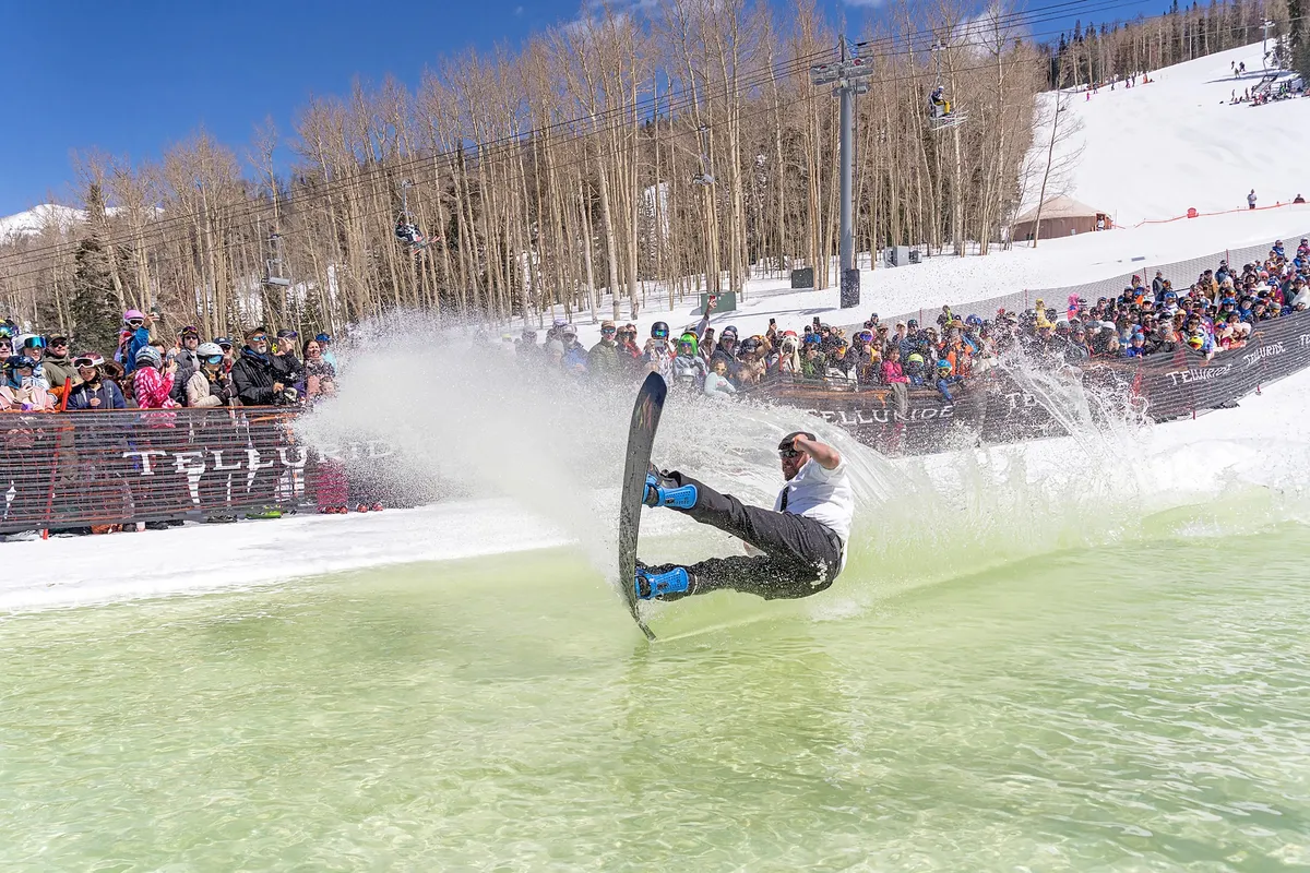 An energetic scene of a snowboarder splashing into a pool, with a lively crowd of spectators and trees in the background. The image conveys excitement and the festive spirit of local events.