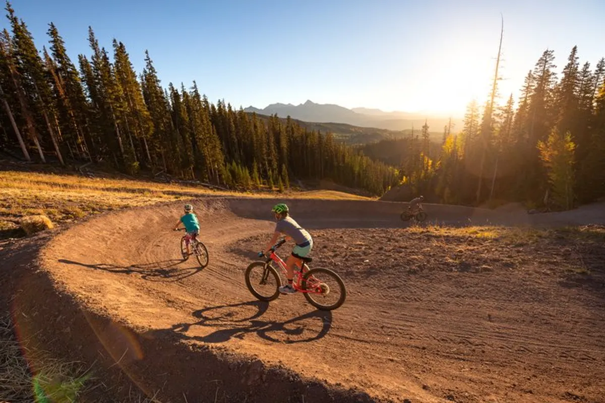 Cyclists navigate thrilling mountain biking trails surrounded by a lush forest, with the sun casting a warm glow over the scene. The energy and terrain highlight Telluride’s renowned hiking and biking access.
