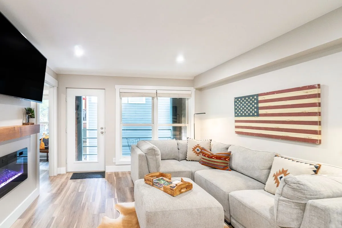 The image showcases a stylish living area with a plush sofa, a striking American flag artwork as a focal point, and a sleek electric fireplace. The space exudes warmth and comfort, perfect for relaxing, socializing, or cozying up for a movie night after a day on the slopes. The ambiance is both modern and inviting, blending contemporary mountain chic with a welcoming, homey feel.