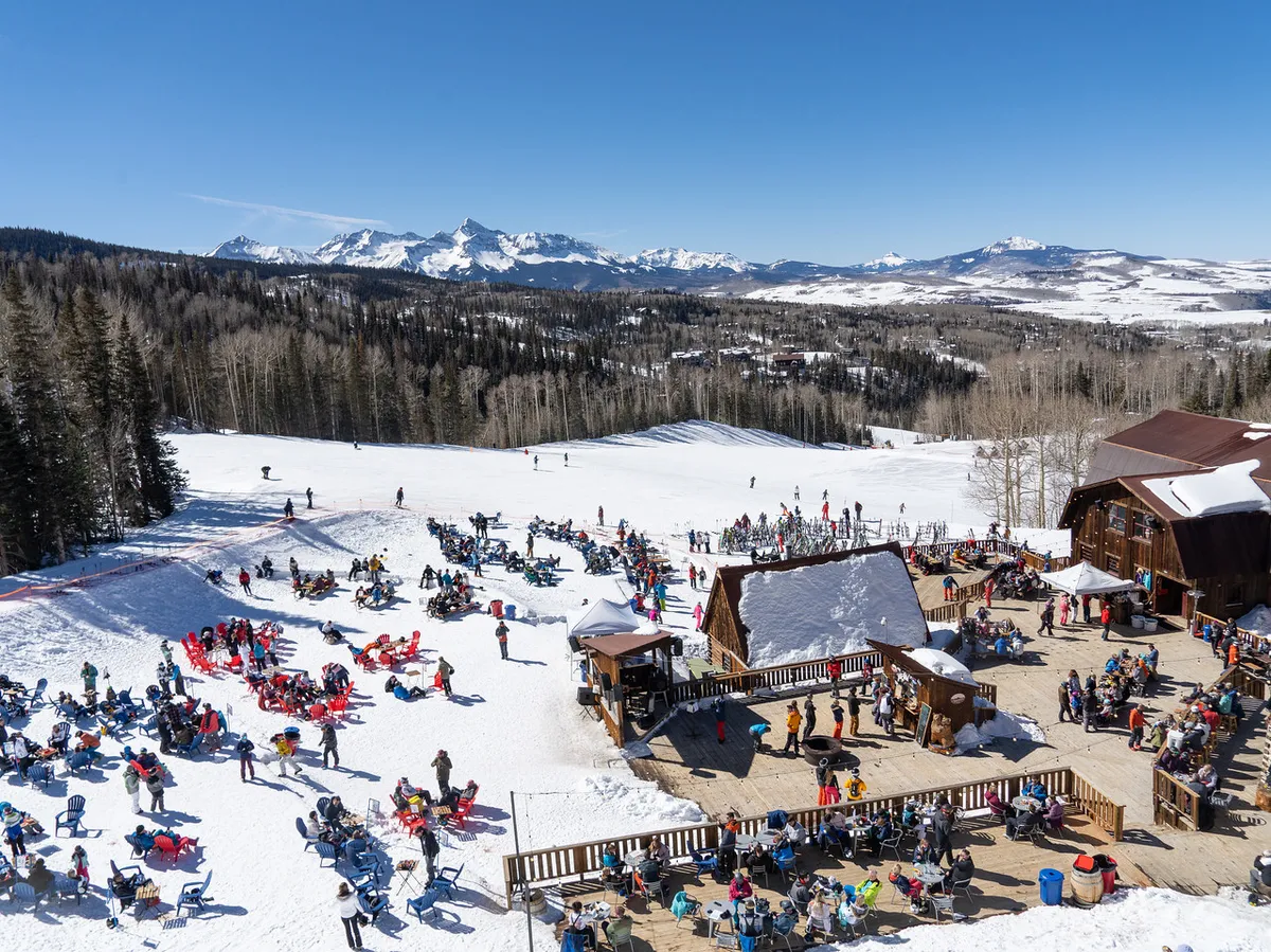 Skiers descend snow-covered slopes with a lively outdoor seating area in the foreground and majestic mountains in the background. The image radiates energy and social excitement after a day on the slopes.