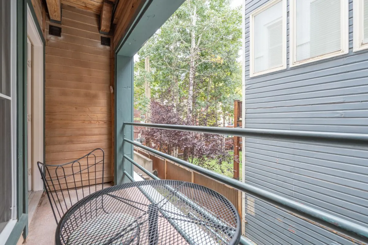 A cozy outdoor patio setting with a metal table and woven chair overlooking lush green trees—a perfect spot for morning coffee or a quiet evening retreat.