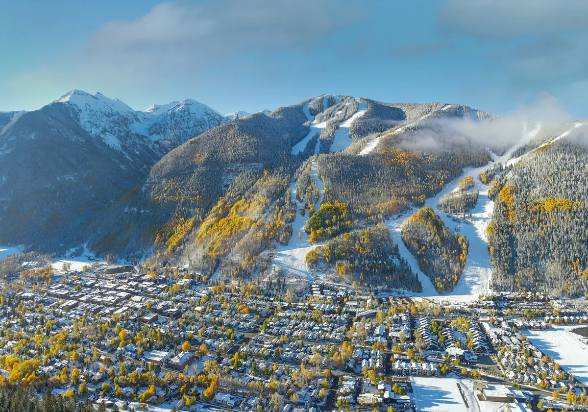 A panoramic view of snow-covered mountains and ski slopes, autumn trees, and the quaint town nestled below. The image conveys both adventure and tranquility, with Telluride’s scenic landscape front and center.