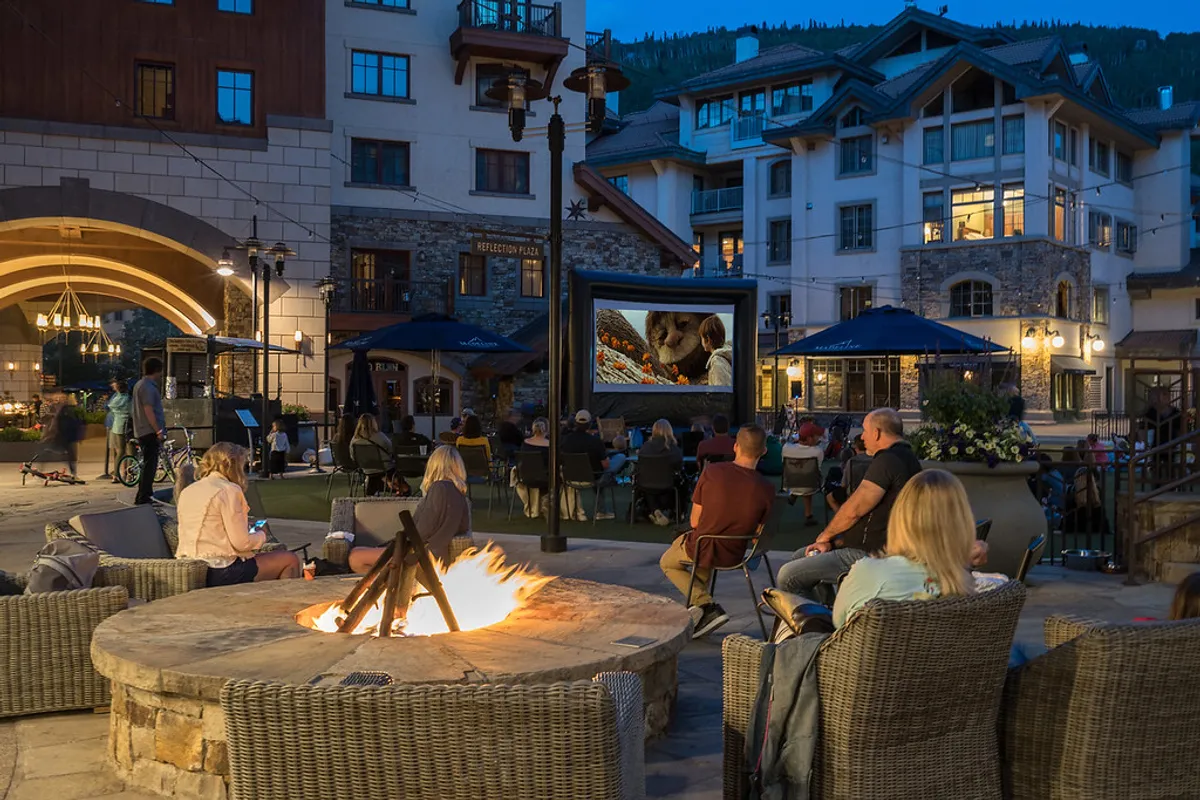 A lively exterior scene with people gathered around a fire pit, an outdoor screen displaying a movie, and a cozy, inviting ambience. The setting is festive, social, and central, suggesting walkable access and group gatherings.