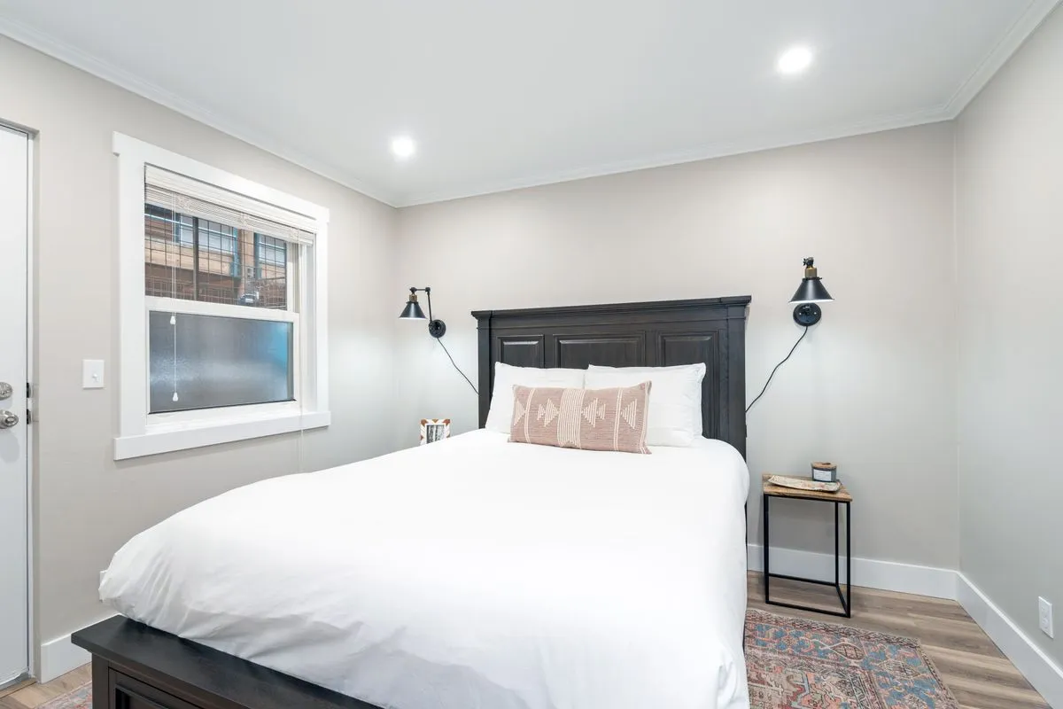 A welcoming bedroom scene with a comfortable bed accented by a decorative pillow, nightstands, and a window providing gentle natural light. The setting feels cozy and inviting, perfect for a restful night's sleep or a quiet morning with coffee.