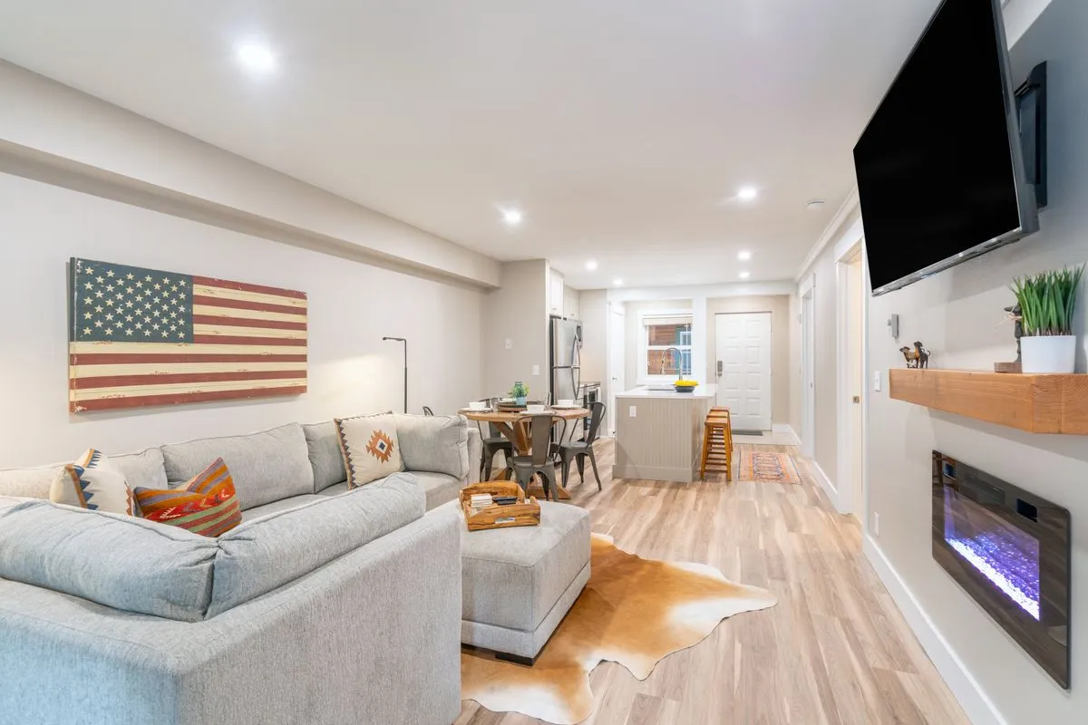 A spacious, warmly lit living room anchored by a plush modular sectional sofa facing a modern electric fireplace. American flag artwork adds a unique decorative touch above the fireplace. The open-concept layout includes a glimpse of a modern dining table, suggesting ample space for gatherings. The ambiance is cozy, inviting, and designed for both relaxing and socializing, with a Smart TV mounted above the fireplace for entertainment.
