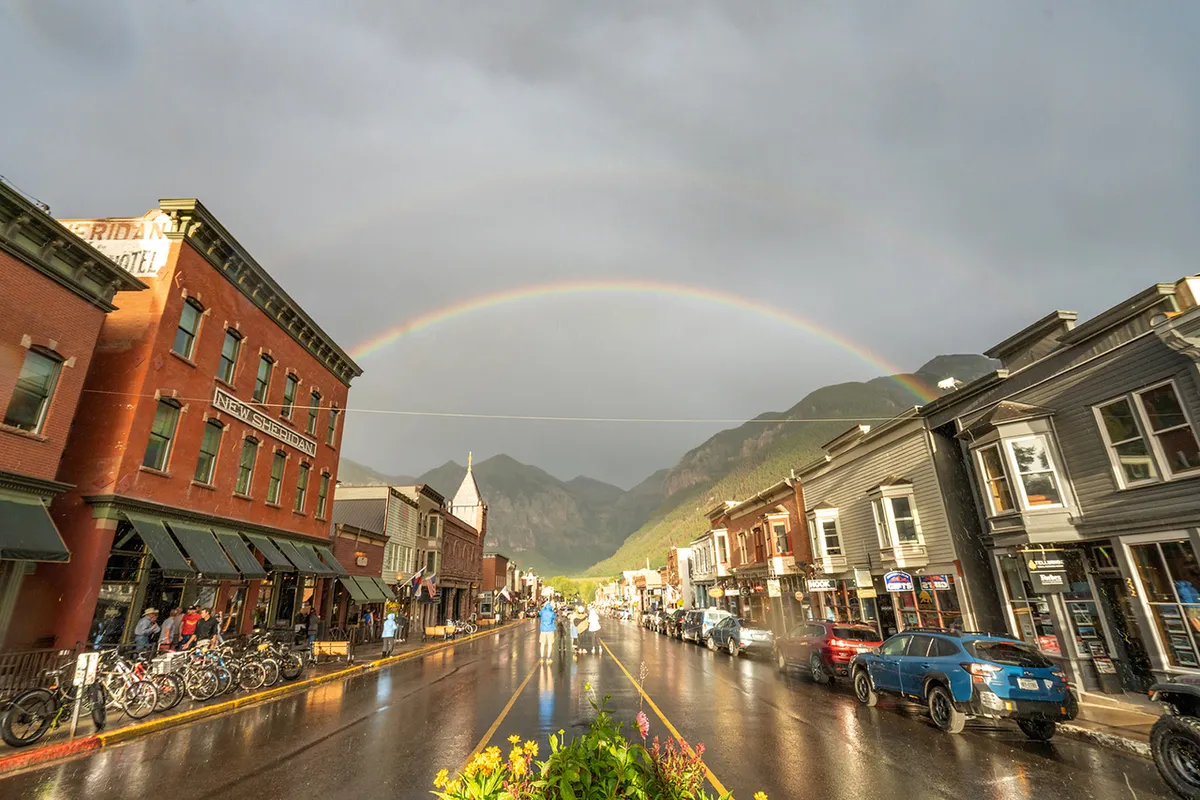 A cheerful Telluride street scene featuring historic buildings, a vibrant rainbow arching over a wet street, and mountains in the distance. The area is lively, evoking the charm and activity of Main Street’s shops, dining, and family outings.