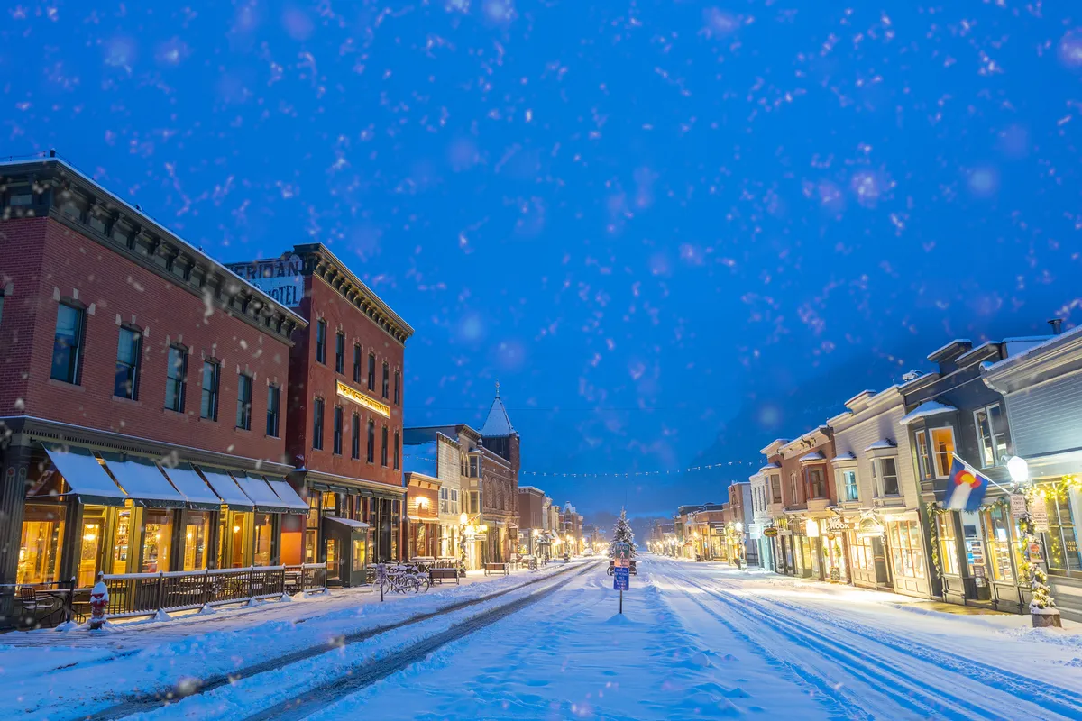 A snow-covered street in Telluride lined with historic buildings illuminated by decorative lights. The scene suggests an inviting, festive atmosphere perfect for winter strolls, holiday shopping, and exploring the town's charm.