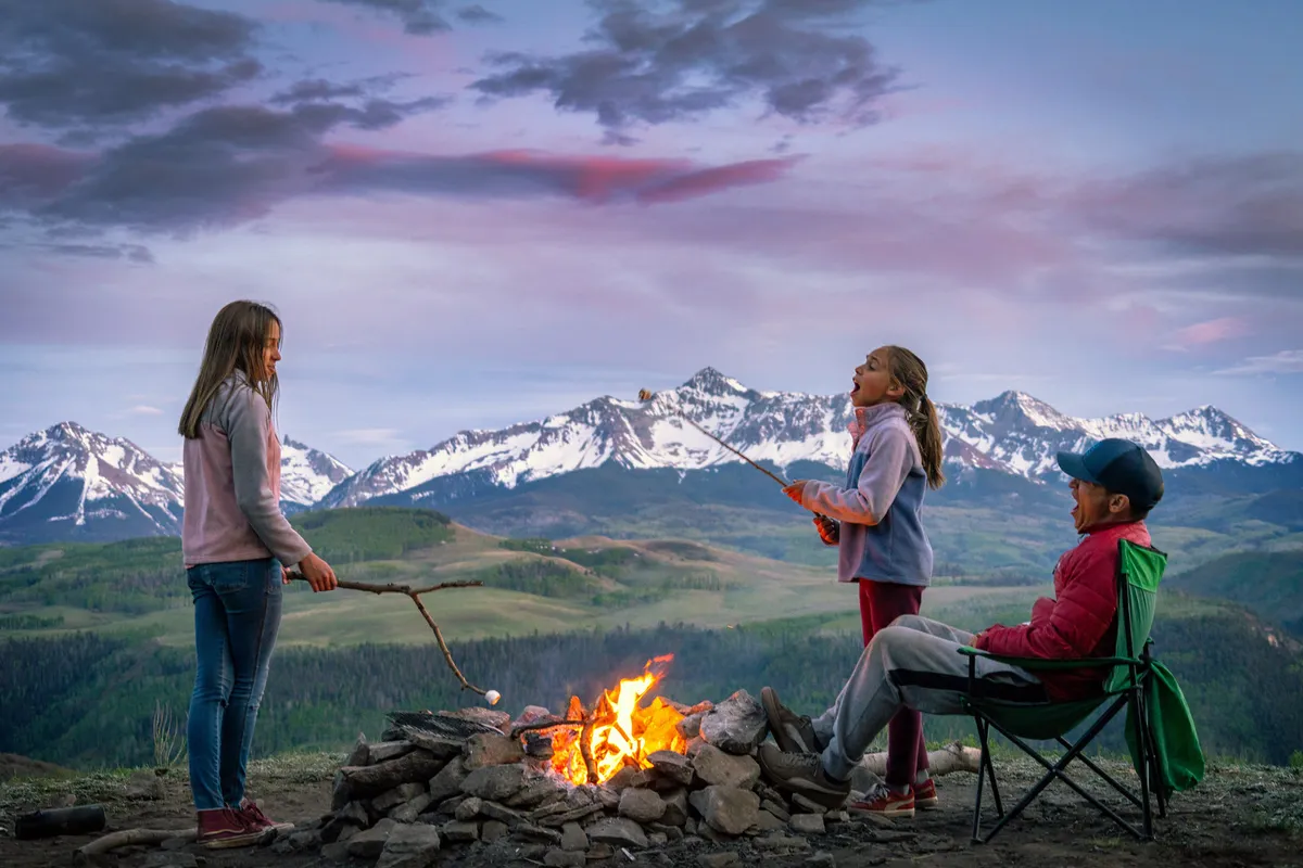 A joyful family scene outdoors: children roast marshmallows around a campfire, with mountains providing a stunning backdrop. The warm light and happy faces capture the magic of shared family experiences in nature.