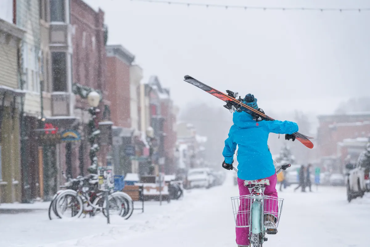 A bustling snow-covered street scene with a person riding a bike, skis visible, vibrant shopfronts, and a sense of active, wintertime community life.