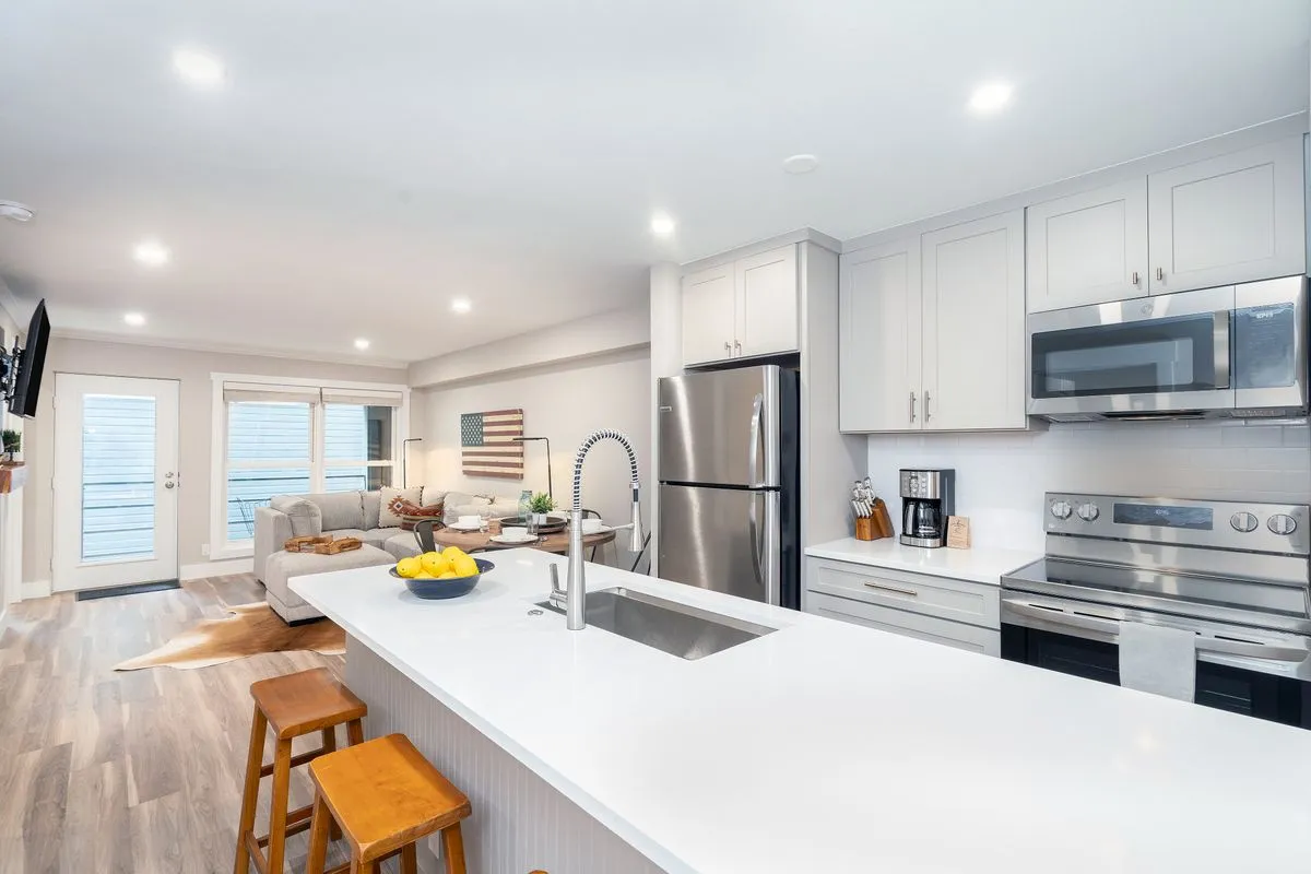 This image features a kitchen island with bar stools, gleaming stainless steel appliances, and a view into the adjoining living area with a cozy sofa. The space exudes warmth and is set up for cooking, entertaining, and family gatherings.